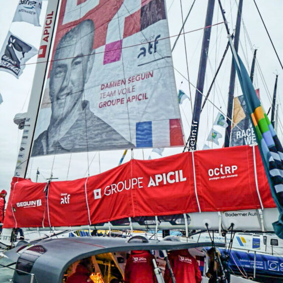 Le bateau de Damien Seguin en mer, coque rouge, la voile rouge et blanche avec le portrait du navigateur, mention Apicil sponsoring