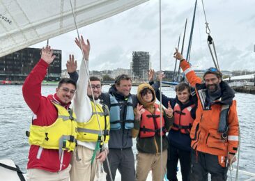 Photo d'un groupe de jeunes bénéficiaires de l'OCIRP, sur le bateau imoca partage, avec François Guiffant, souriants et levant les bras en l'air