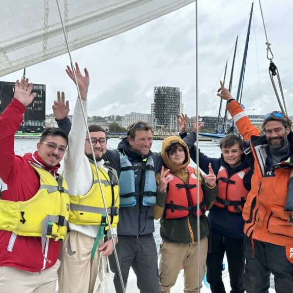Photo d'un groupe de jeunes bénéficiaires de l'OCIRP, sur le bateau imoca partage, avec François Guiffant, souriants et levant les bras en l'air